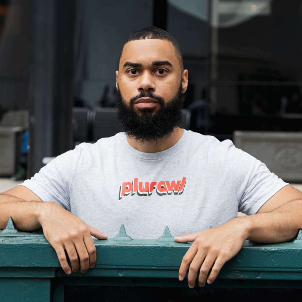 Headshot of Pabel Martinez, an Afro-Latino with a low black haircut, thick black facial hair, and grey crew neck t-shirt with the word “plurawl” printed in bright orange.