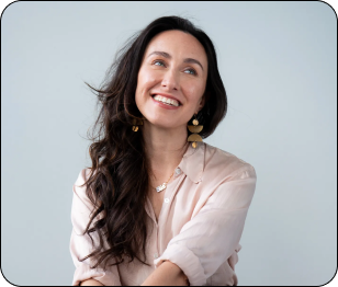 Headshot of Natalia Villalobos, a mixed-race White-presenting cisgender woman with long wavy brown hair, gold dangly earrings, pendant necklace, button-up blush shirt, and black pants sitting on a stool and smiling with her eyes away from the camera with a light gray background