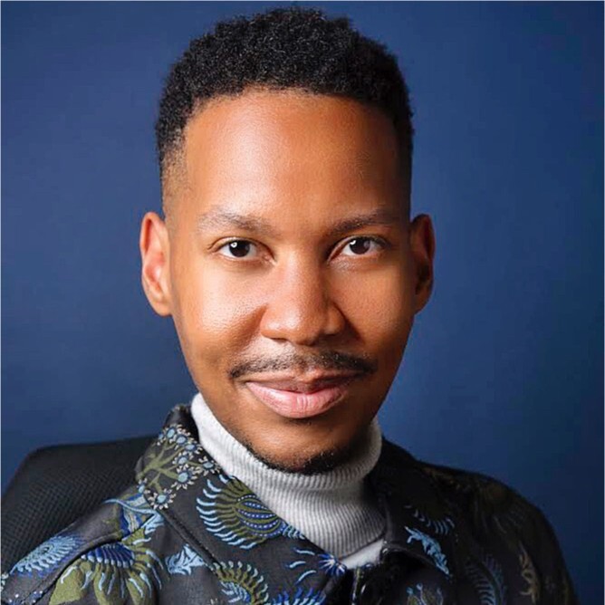 Headshot of Eddie Ndopu, a Black man with short curly black hair and mustache, light grey turtleneck, and black button-down with abstract green and blue floral pattern.
