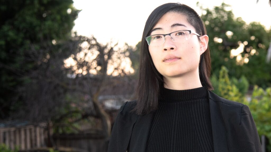 Headshot of Lily Zheng, a Chinese American nonbinary person with dark brown asymmetric hair that is half shaved, glasses, a black shirt, and a black jacket with some trees on the back.