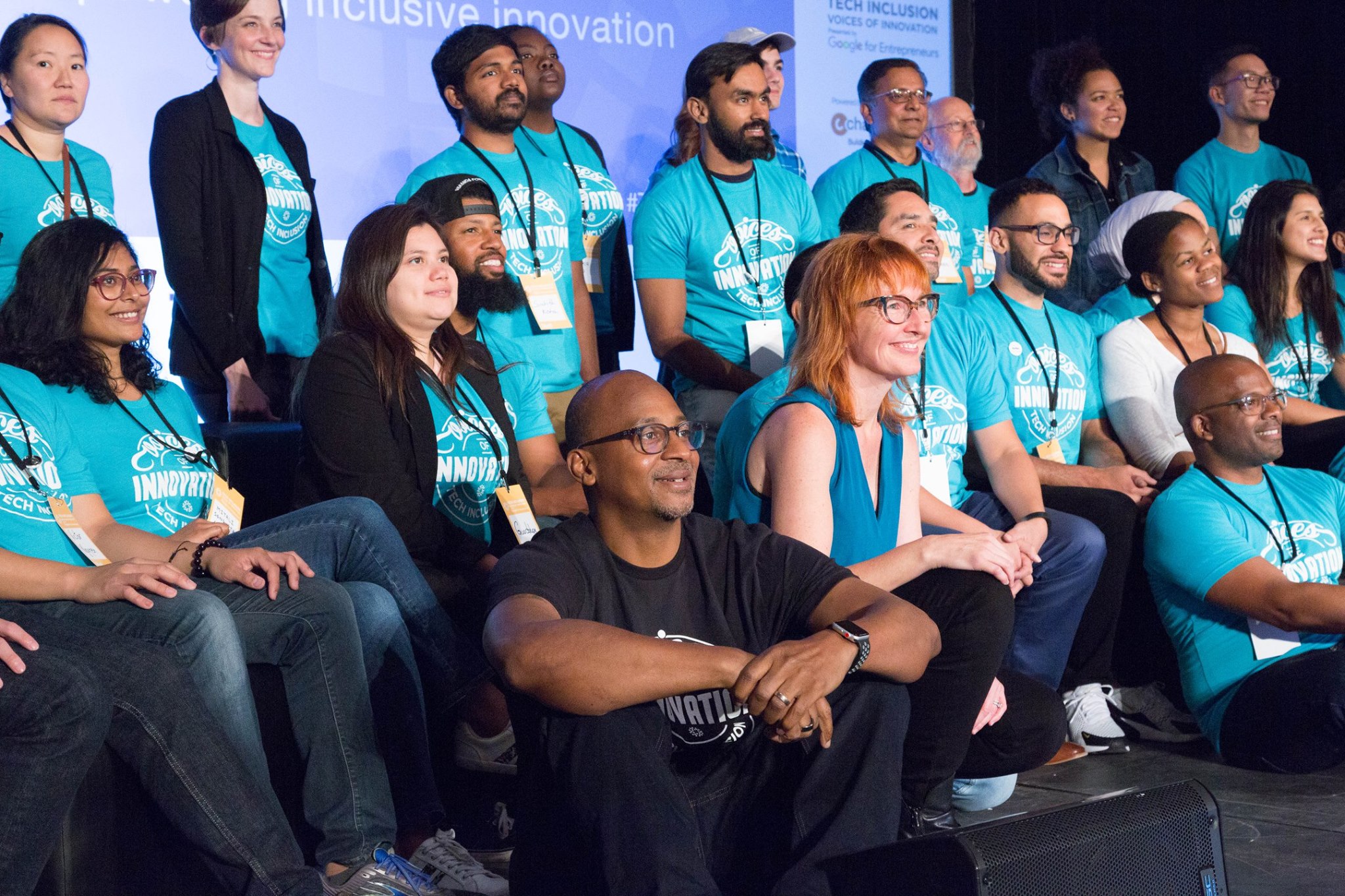 A group of several Tech Inclusion workers in a group photo on stage wearing matching blue t-shirts