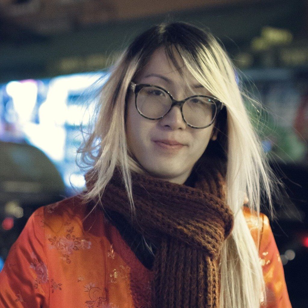 Headshot of Madelena Mak, an Asian trans person, blonde long hair, wearing orange floral coat and brown scarf and glasses