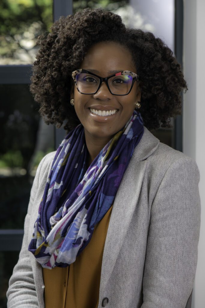 Headshot of Dr. Adia Gooden, Black women wearing a grey coat, orange blouse, blue and purple scarf, leopard glasses, and golden earrings