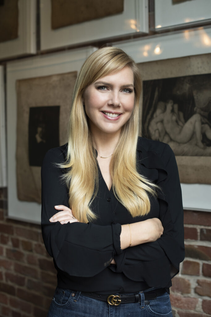 Headshot of Katelin Holloway, a White Mom with long blonde hair and a black blouse, in front of a brick wall with some paintings.