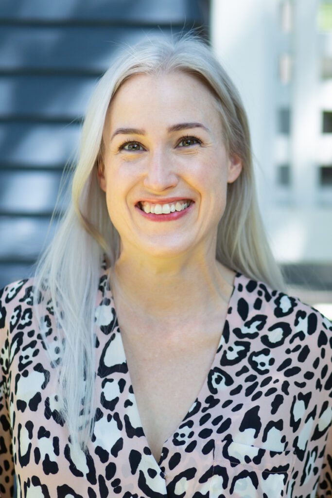 Headshot of Kt McBratney, a White nonbinary person with long platinum hair and a pink animal print blouse.