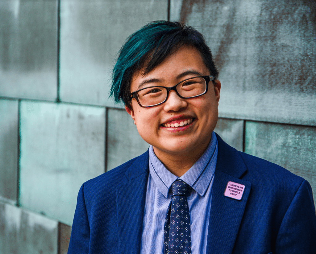 Headshot of Lydia X. Z. Brown, an East Asian person with short black and teal hair, glasses, a dark blue suit, a light blue button-down, a diamond-patterned tie, and a square pin on their suit that reads: THERE IS NO WRONG WAY TO HAVE A BODY, smiling at the camera in front of a grey brick wall.