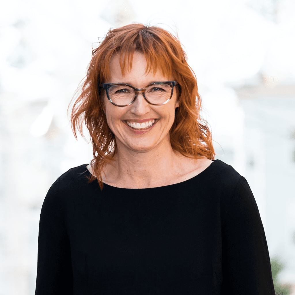 Professional photo of Melinda, a White woman with red hair, glasses, and black long-sleeve shirt looking at the camera, smiling