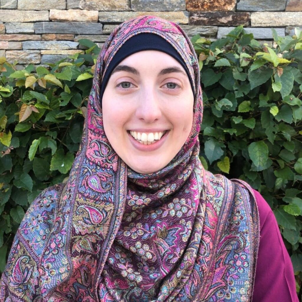 Headshot of Rahimeh Ramezany, a pale-skinned Muslim woman wearing a multi-colored religious headscarf with pinks, blues, and purples and a fuchsia blouse, smiling at the camera with green bushes at the back.