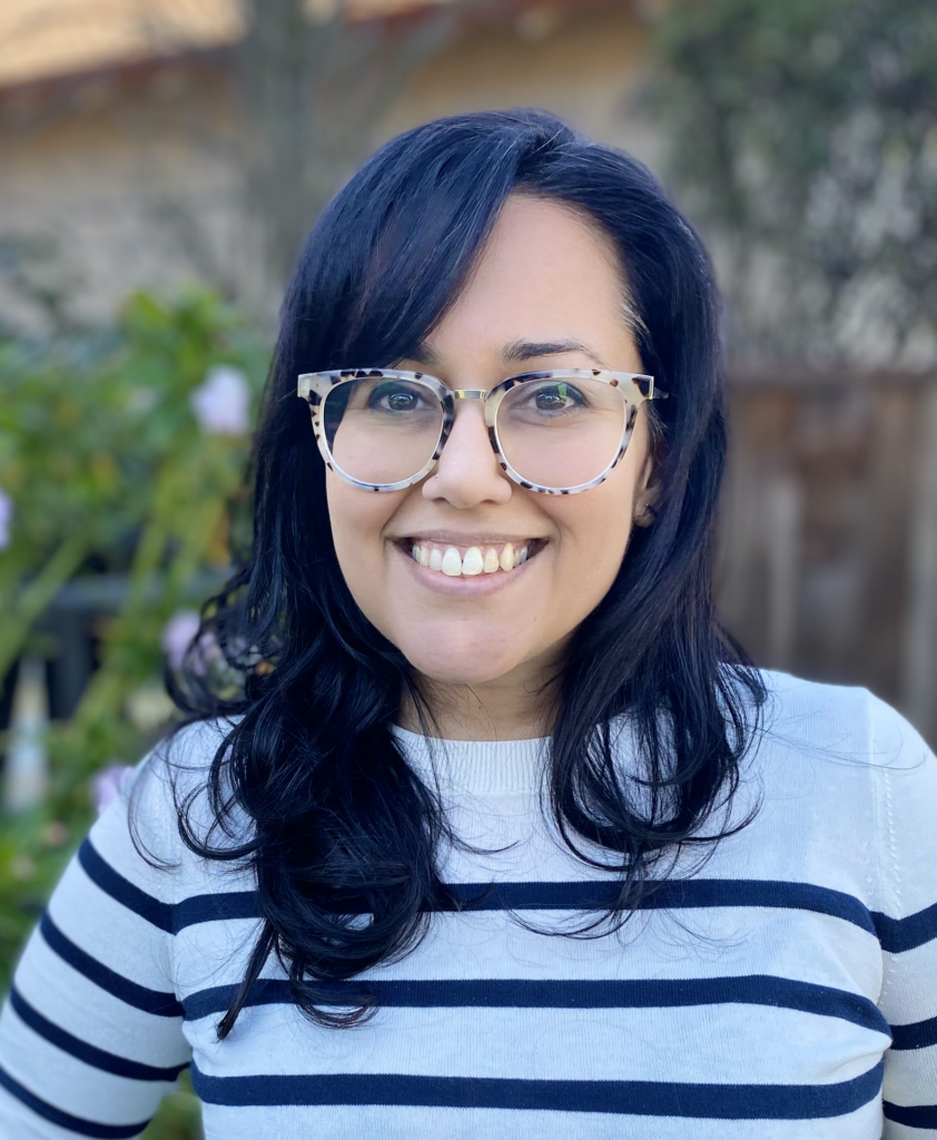 Headshot of Nisha Anand, a South Asian female with long wavy black hair, brown eyes, glasses, and a striped black and white long sleeve