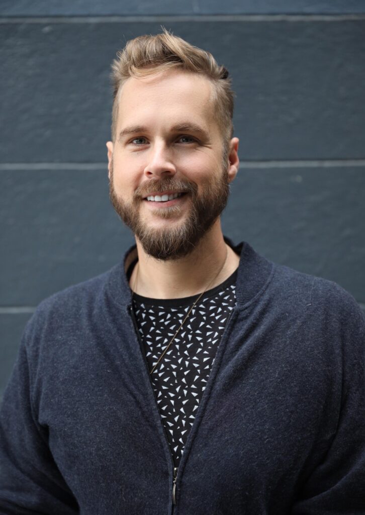 Headshot of John Marble, a White man with blond hair and a brown beard, a black t-shirt with a small white triangle pattern, and a navy zip-up jacket, smiling at the camera in front of a grey wall