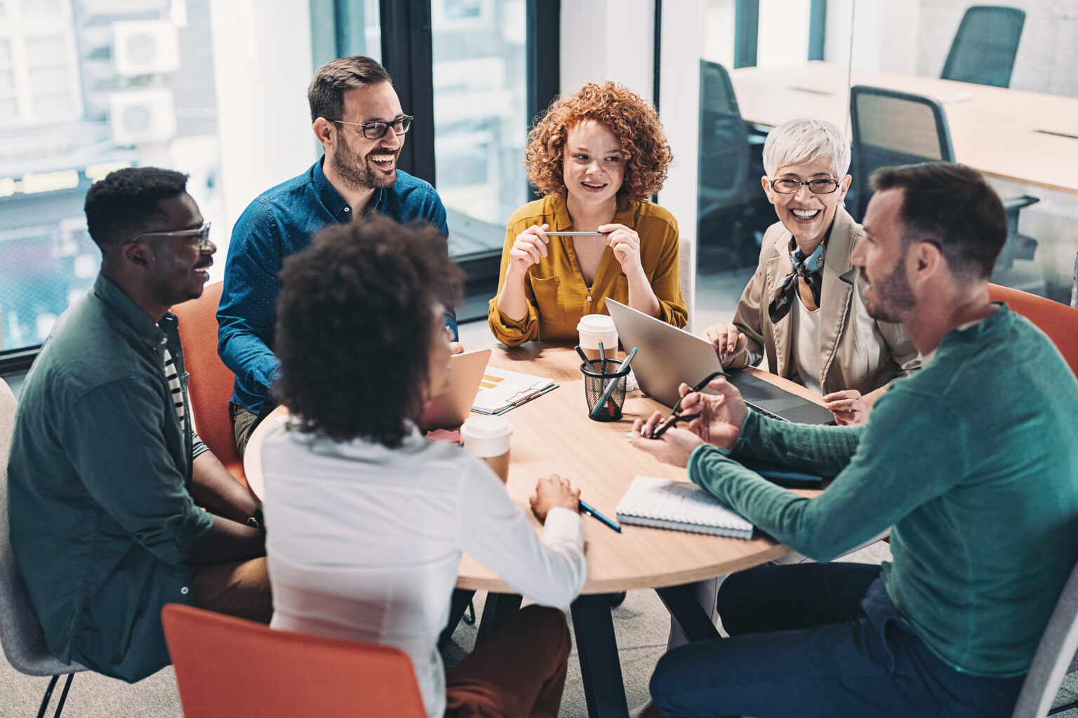 Image of a diverse group of people sitting around a table and talking with notepads and laptops