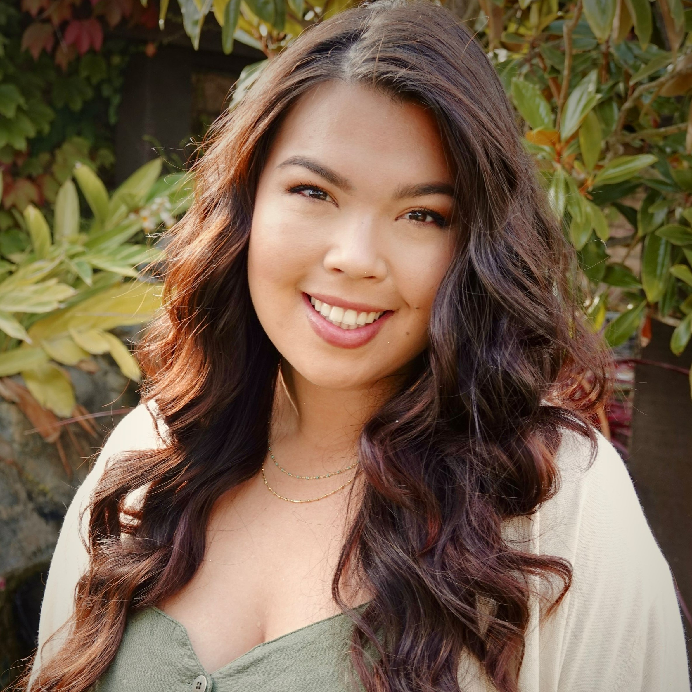 Headshot of Christina, a biracial Asian and White woman with curled long brown hair, an olive green top, and cream-colored cardigan smiling at the camera outdoors with a large plant in the background.