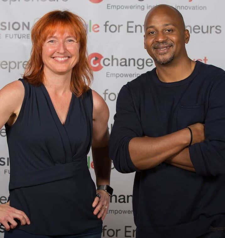 Photo of Melinda, a White woman with red hair and dark blue sleeveless shirt, and Wayne, a bald Black man with a goatee and dark blue long-sleeve shirt, smiling at the camera and standing in front of a step-and-repeat backdrop