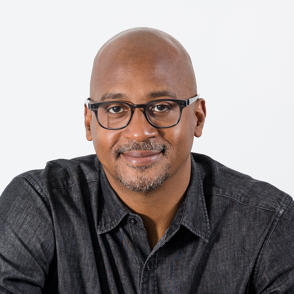 Headshot of Wayne Sutton, a bald Black man with a trimmed goatee, glasses, and a charcoal button-down looking at the camera with a white background
