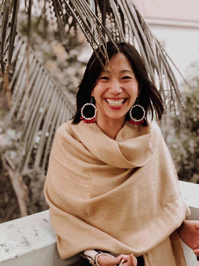 Headshot of Tiffany, an Asian woman with shoulder-length brown hair, red and white beaded hoops, a beige shawl. and a brace on her right wrist smiling at the camera and leaning against a low white wall with the fronds of a palm tree brushing against her head.