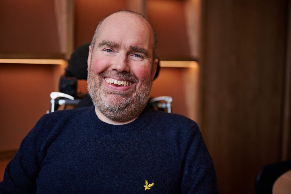 Headshot of Toby Mildon, a White man sitting in an electric wheelchair with low haircut, salt and pepper beard, and navy blue long-sleeve shirt smiling at the camera with a blurred wood wall in the background.