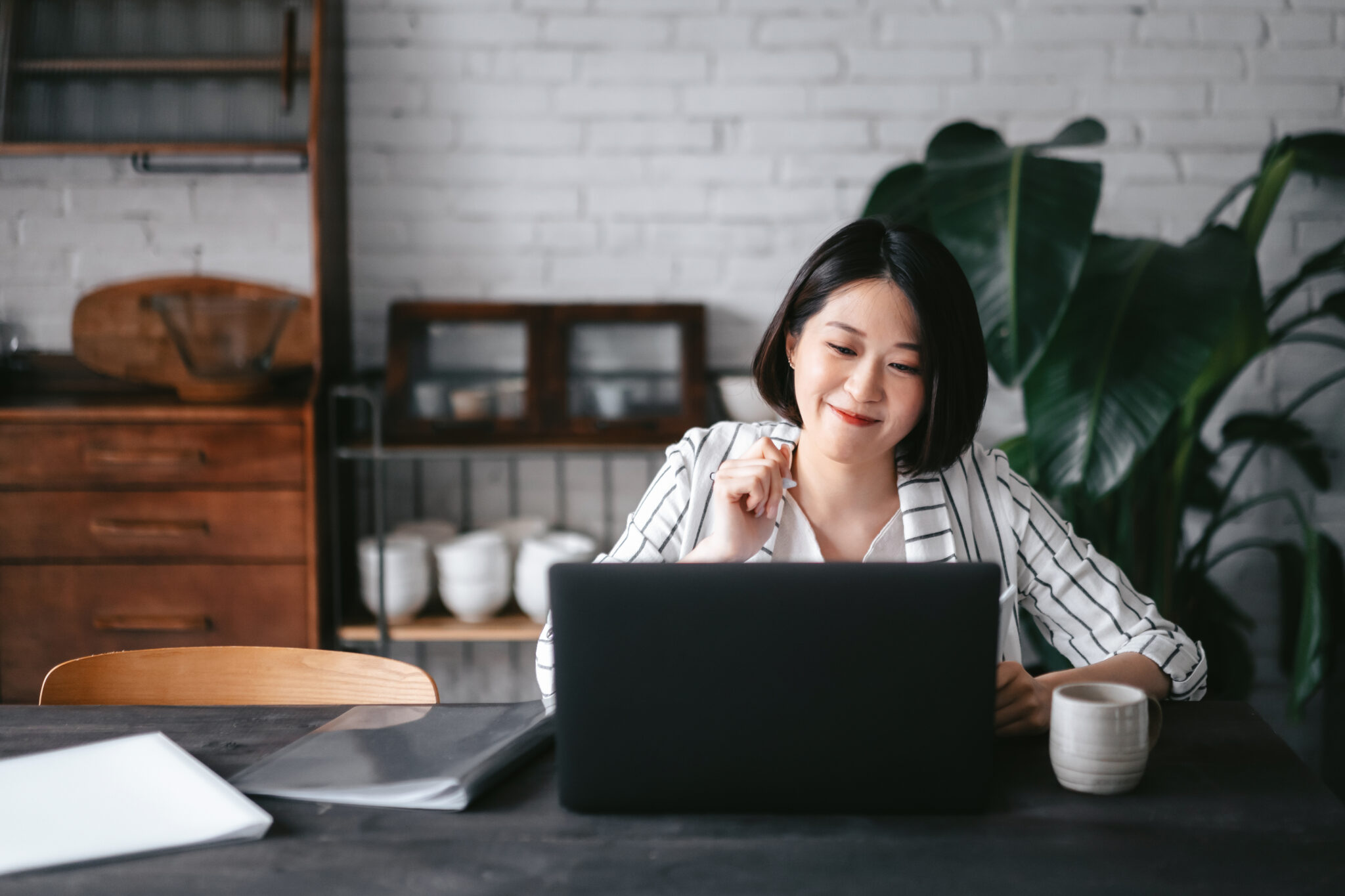 Asian feminine-presenting person sitting at a table in front of their laptop and a coffee mug with a white brick wall, wood cabinets, and a large plant in the background