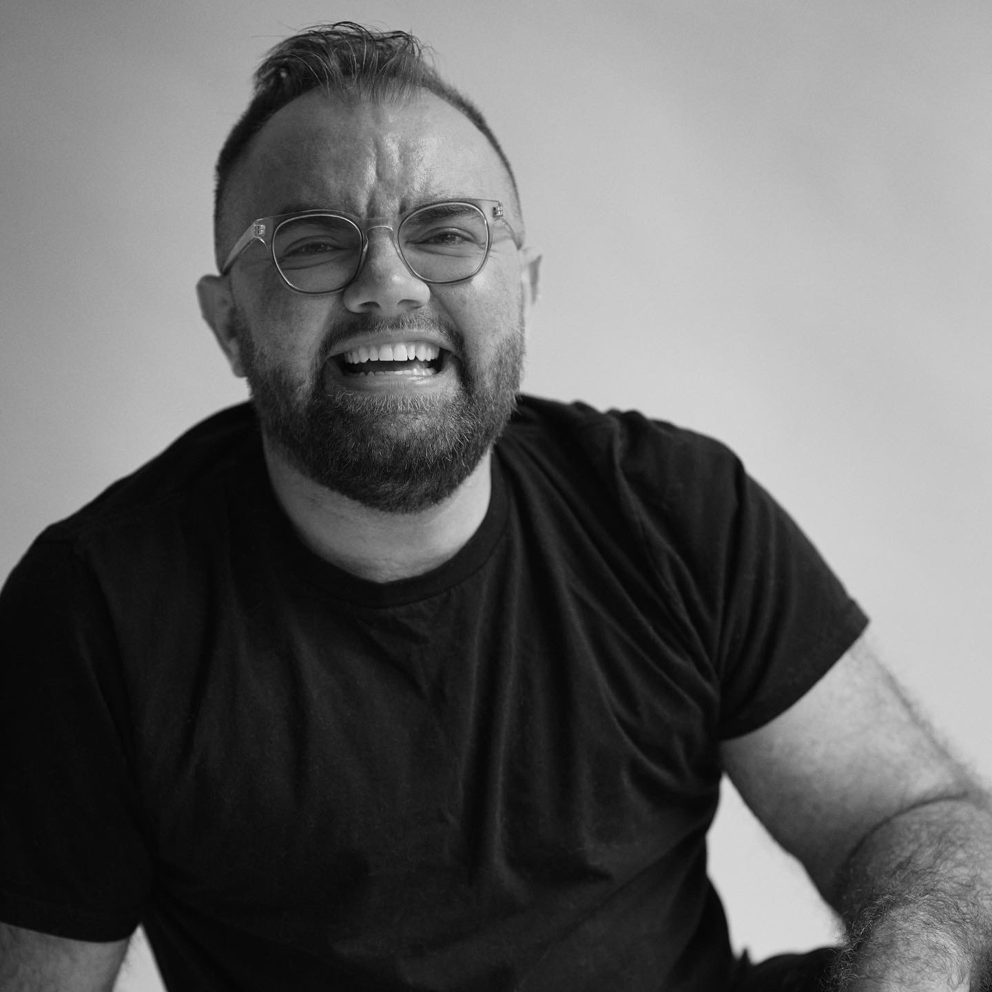 Black and white photo of a nonbinary person with short hair and facial hair, glasses, and a dark t-shirt smiling at the camera in front of a lighter background.