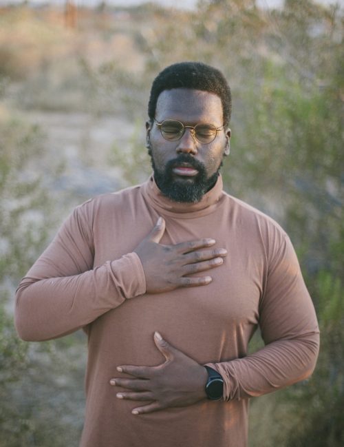 Headshot of Aaron Johnson, an African Heritage man with short black afro hair, black goatee and beard, tan turtleneck, glasses, and a black watch with his eyes closed and one hand each on his chest and belly in front of a blurry outdoor plant-filled background.