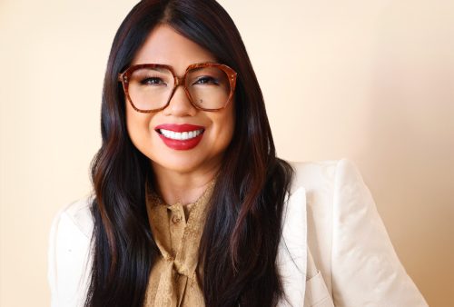 Headshot of Marissa Andrada, an Asian Mixed Race Woman with long brown hair, tortoiseshell glasses, gold blouse, and white blazer smiling at the camera in front of a light beige wall.