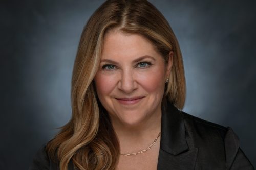 Headshot of Megan Carle, a White woman with long dark blonde hair, rhinestone stud earrings, a gold chain necklace, and black blazer looking at the camera in front of a grey backdrop