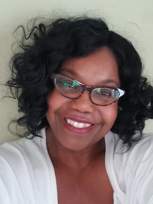 Headshot of Cory, a Black Indigenous woman with short curly black hair, glasses, and a white top smiling at the camera in front of an off-white wall